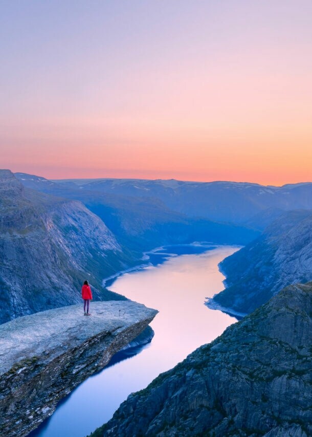 Bergklippe Trolltunga mit Person in roter Jacke, die auf dem Felsvorsprung über einem tiefen Fjord steht, umgeben von Bergen im Abendlicht.