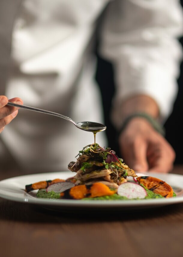 Die Hände eines Kochs, der Soße aus dem Löffel auf eine Mahlzeit auf einem weißen Teller gießt.