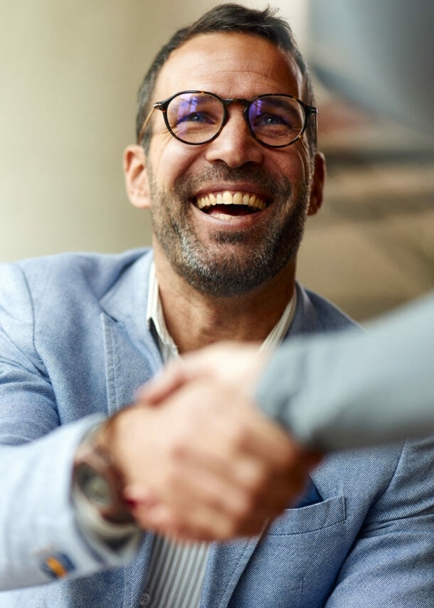 Ein lächelnder Mann im hellblauen Sakko schüttelt einer Person die Hand.