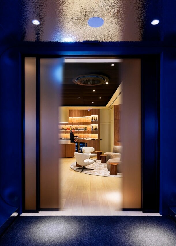 Blick durch einen dunklen, blau beleuchteten Gang mit vier rechteckigen Bildschirmen an den Seiten auf einen hellen, modernen Raum mit Sesseln und Regalen.