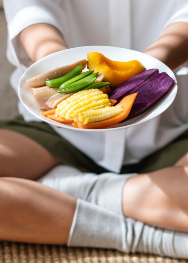 Person sitzt im Schneidersitz und hält einen Teller mit buntem, geschnittenem Gemüse, darunter Mais, Süßkartoffel und grüne Bohnen.