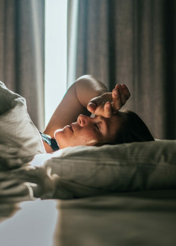 Eine Person liegt auf einem Bett, mit einem weißen Bettlaken und Kissen. Licht strömt durch ein Fenster, das von Vorhängen umgeben ist.