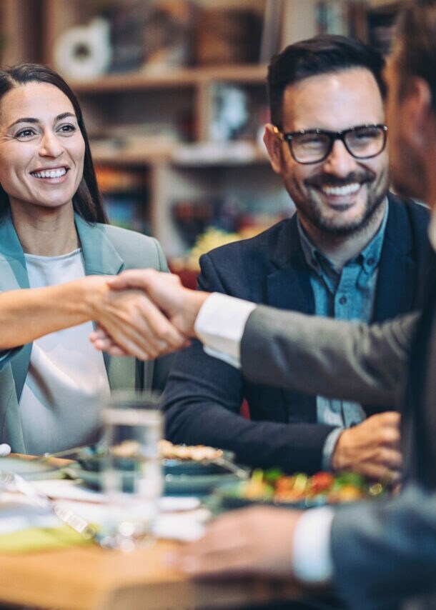 Eine lächelnde, elegant gekleidete Frau im Blazer und ein Mann im Anzug geben sich die Hand an einem Tisch mit zwei weiteren Personen.