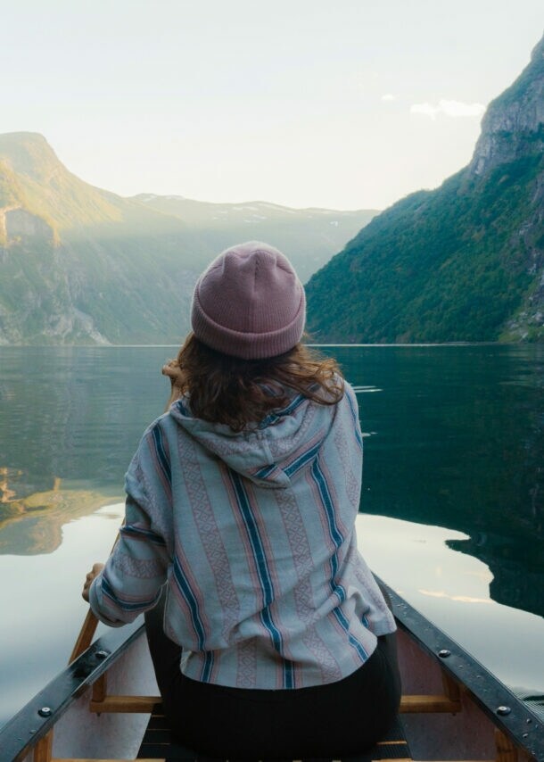 Person mit rosa Mütze und gestreiftem Pullover sitzt in einem Kanu auf ruhigem Wasser, umgeben von hohen, bewaldeten norwegischen Fjordwänden mit Wasserfall.