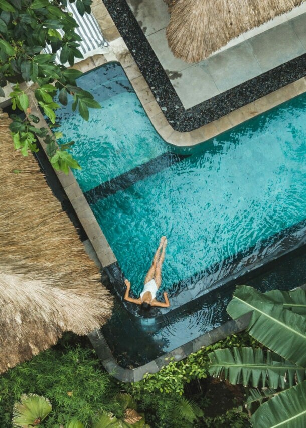 Eine Person schwimmt in einem blauen Pool, umgeben von tropischen Pflanzen und einem Strohdach. Der Blick ist von oben.