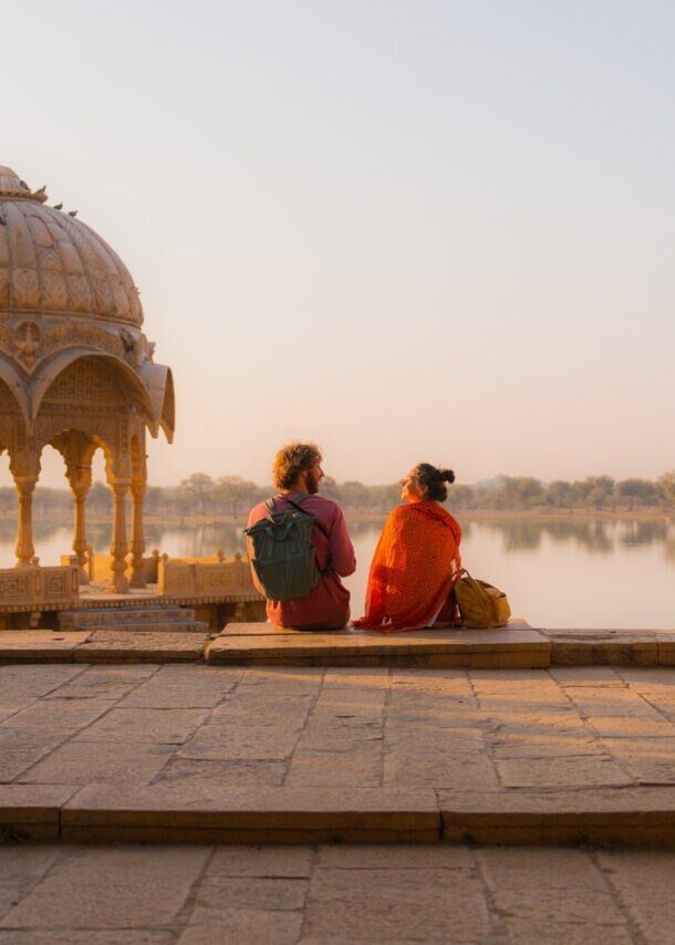 Zwei Personen mit Rucksäcken sitzen in einer buddhistischen Tempelanlage an einem See im weichen Sonnenlicht.