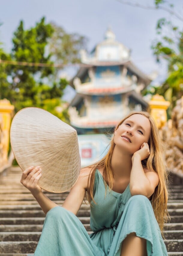 Eine Frau sitzt lächelnd auf einer Treppe. Sie schwenkt einen traditionellen vietnamesischen Hut. Im Hintergrund sind ein Tempel und goldene Statuen zu sehen.