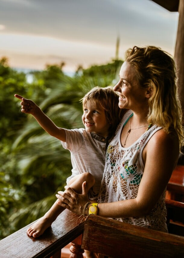 Eine Frau hat einen kleinen Jungen im Arm, beide blicken von einer Holzterrasse in den Dschungel von Thailand, hinter ihnen das Meer.