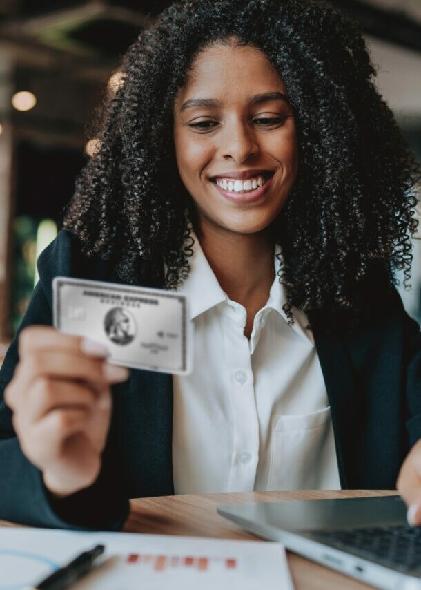 Eine lächelnde Frau bedient mit einer Hand einen Laptop und hält in der anderen Hand eine silberne Kreditkarte von American Express.