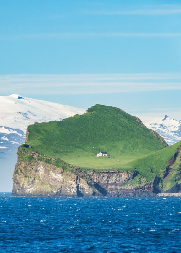 Blick auf die grüne isländische Insel Ellidaey mit einer einsamen Jagdhütte darauf und dem, unter einem Gletscher liegenden Vulkan Eyjafjallajökull im Hintergrund.