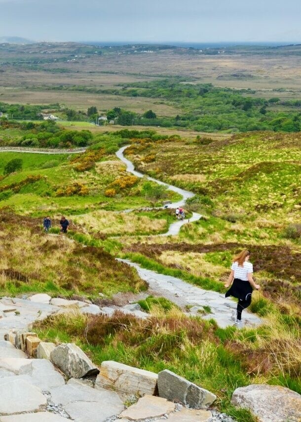Heide- und Wiesenlandschaft, durch die sich ein steiniger Weg mit wandernden Personen darauf schlängelt, im Hintergrund ein Berg und Wasser.