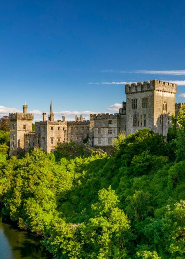 Luftaufnahme von Lismore Castle in Waterford, umgeben von dichtem Grün und einem Fluss mit einer Steinbrücke im Vordergrund.