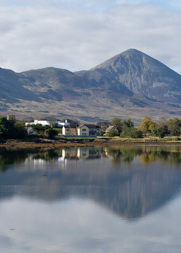 Vor einem großen Gewässer stehen kleine Häuser und Bäume, in Hintergrund ragt ein großer Berg empor, die Szene spiegelt sich in der Wasseroberfläche.