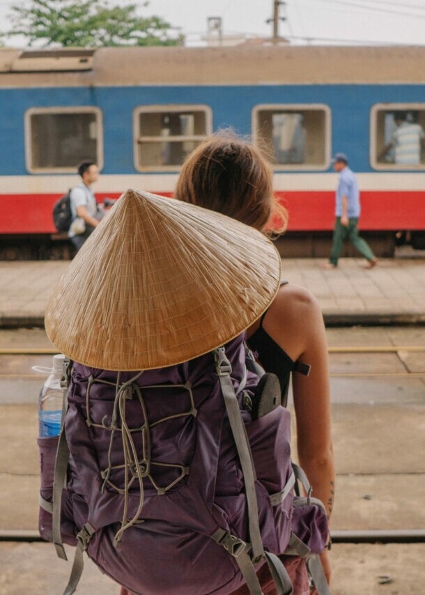 Eine Person mit einem großen, runden Strohhut und einem lila Rucksack steht auf einem Bahnsteig und schaut auf einen Zug. Im Hintergrund sind mehrere Personen und ein Zug zu sehen.