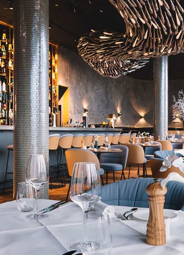 Stilvoller, modern eingerichteter Speisesaal mit Barbereich und eingedeckten Tischen unter einer abstrakten Deckenskulptur.