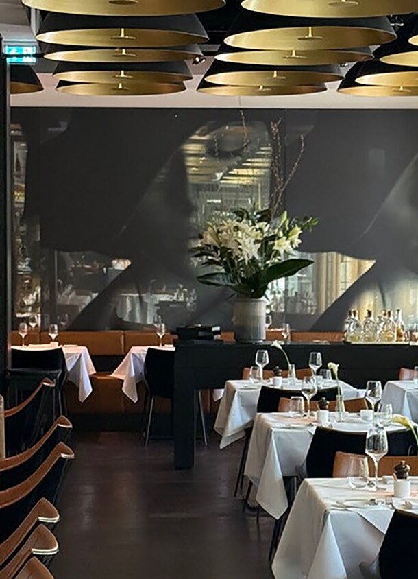 Eleganter Speiseraum mit weiß eingedeckten Tischen und goldfarbenen Deckenlampen vor einem schwarzen Wandbild.