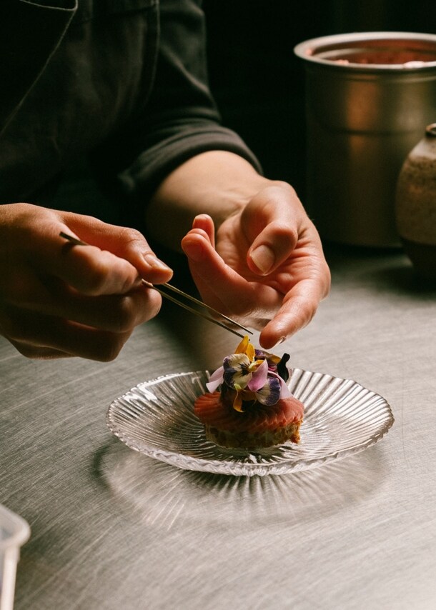 Zwei Hände garnieren eine edle Vorspeise mit Blüten auf einer Küchentheke.