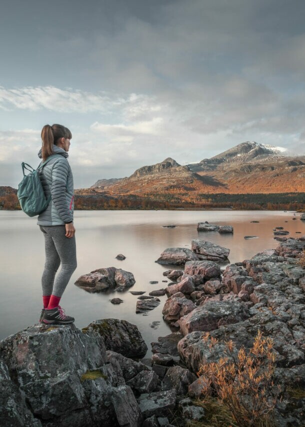 Eine Frau mit Rucksack steht auf einem Stein an einem See in flacher Felslandschaft mit herbstlicher Vegetation.