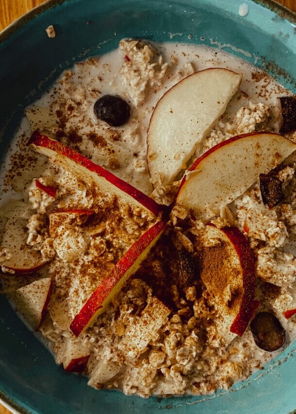 Blaue Schüssel mit Porridge, garniert mit Apfelscheiben, Blaubeeren und Zimt, auf Holztisch mit Glas Wasser und Serviette.