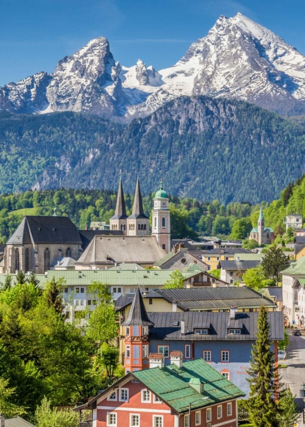 Blick auf eine Stadt mit historischen Gebäuden und einer Kirche vor bewaldeten Hügeln und schneebedeckten Bergen im Hintergrund.