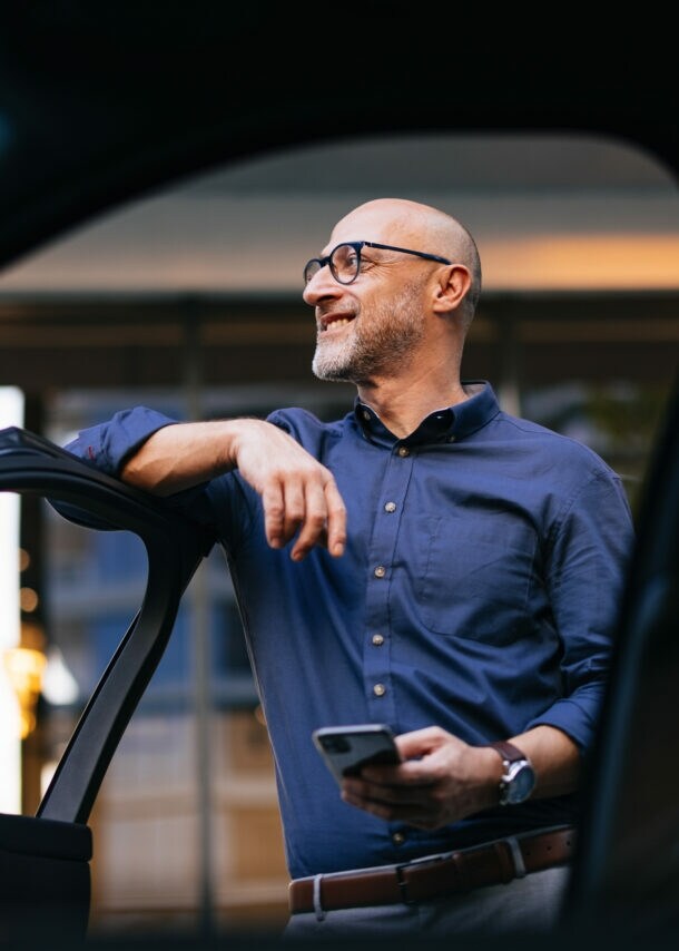 Ein Mann im blauen Hemd und mit einem Smartphone in der Hand lehnt an einer geöffneten Autotür und blickt lächelnd in die Ferne.
