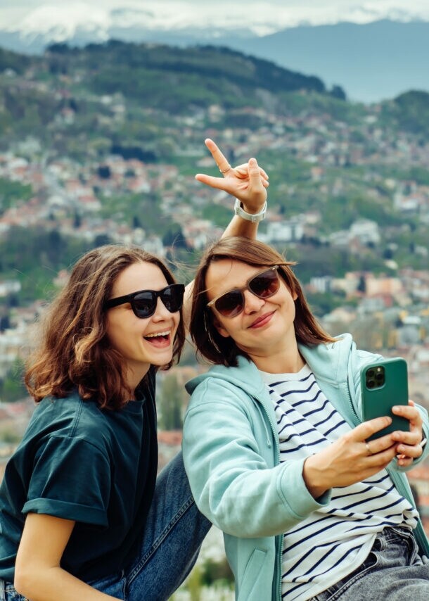 Zwei junge Frauen sitzen auf einer Mauer mit Blick auf eine Stadt, eine macht ein Selfie mit einem Smartphone und zeigt ein Peace-Zeichen.