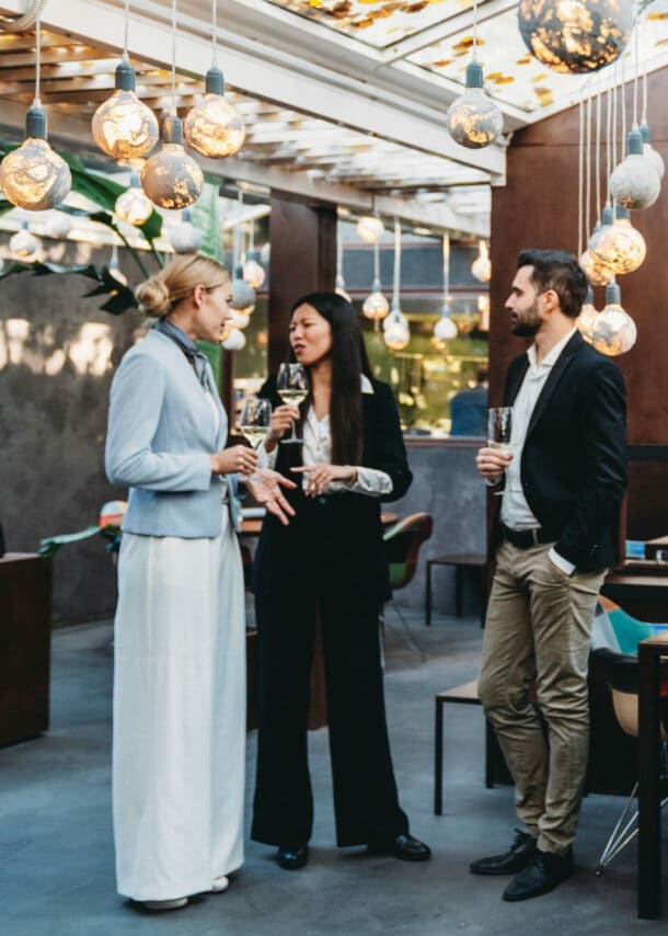 Personen in eleganter Kleidung stehen mit Weingläsern in den Händen beieinander in einem elegant geschmückten Raum.