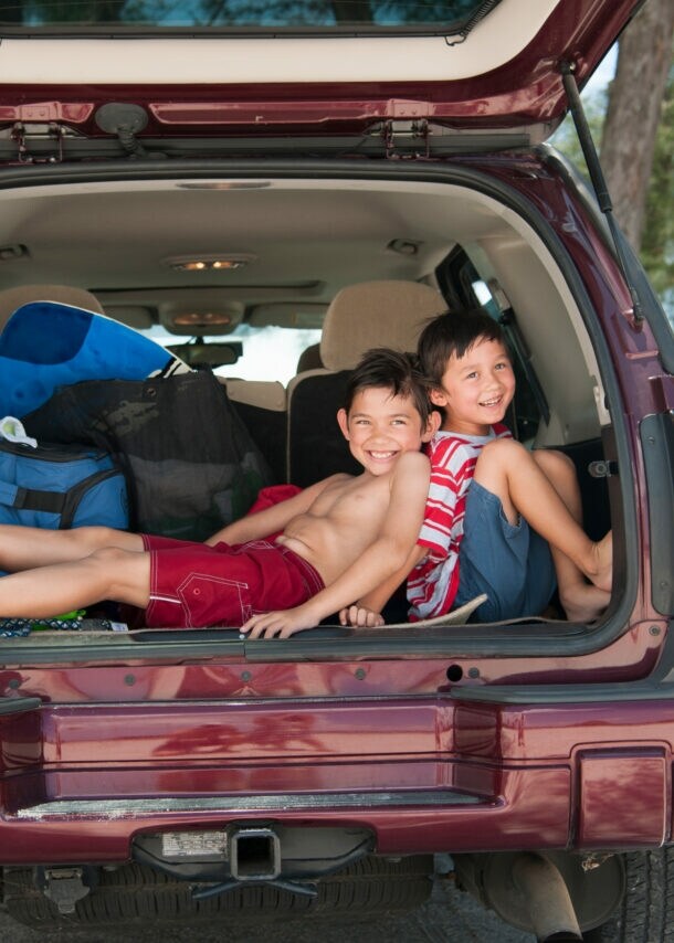 Zwei Jungen in Strandoutfits sitzen fröhlich in einem offenen Kofferraum, hinter ihnen das Meer.