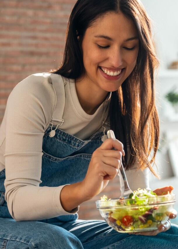 Frau in Jeanslatzhose sitzt auf einem Tisch und isst einen Salat aus einer Glasschüssel mit einer Gabel.