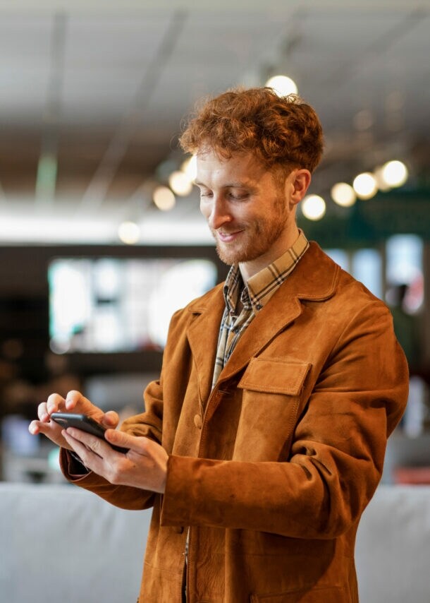Ein junger Mann steht in einem Möbelgeschäft und schaut lächelnd auf das Smartphone in seiner Hand.