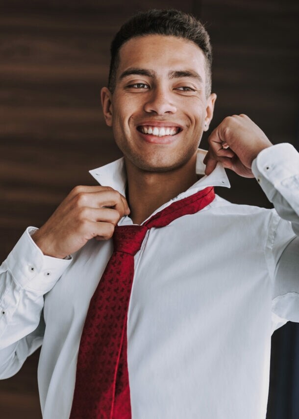 Ein junger Mann im weißen Hemd mit roter Krawatte richtet sich lächelnd den Kragen vor einem Spiegel.