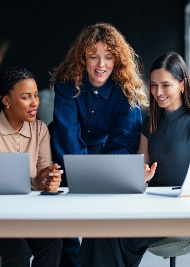 Drei Frauen schauen gemeinsam auf ein Laptop auf einem großen Tisch mit zwei weiteren Laptops in einem modernen Büro.