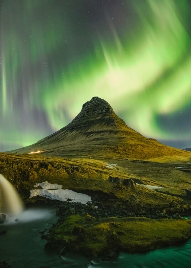 Moosbedeckte Vulkanlandschaft mit Wasserfällen unter Nachthimmel mit grün und lila leuchtenden Polarlichtern.