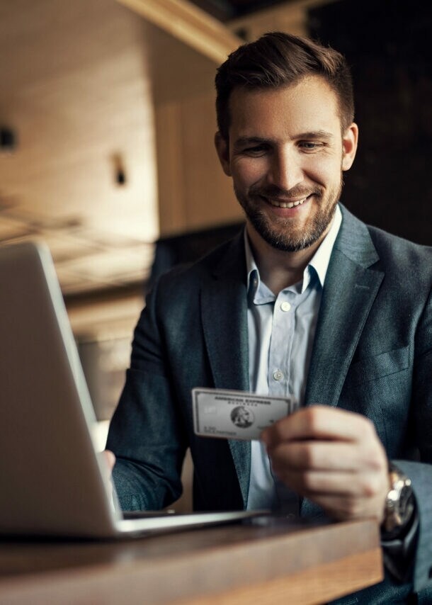 Ein lächelnder Geschäftsmann sitzt an einem Tisch vor einem Laptop und blickt auf eine silberne Platinum Kreditkarte von American Express in seiner Hand.