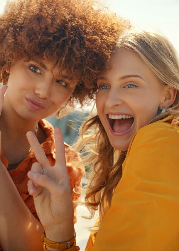 Zwei junge Frauen zeigen Peace-Zeichen mit ihren Fingen, während sie ein Selfie per Smartphone aufnehmen unter freiem Himmel.