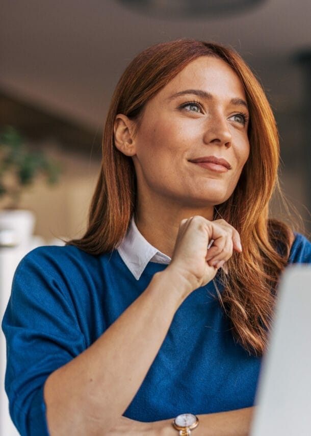 Eine Frau im blauen Pullover sitzt vor einem Laptop und schaut zufrieden lächelnd in ein Büro.