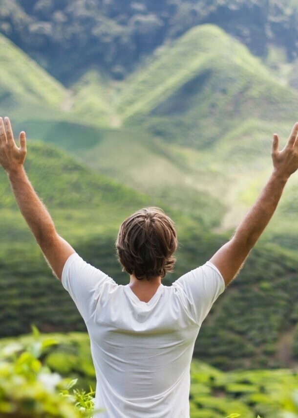 Mann mit weißen T-Shirt steht mit erhobenen Armen vor grünen, bewaldeten Hügeln.