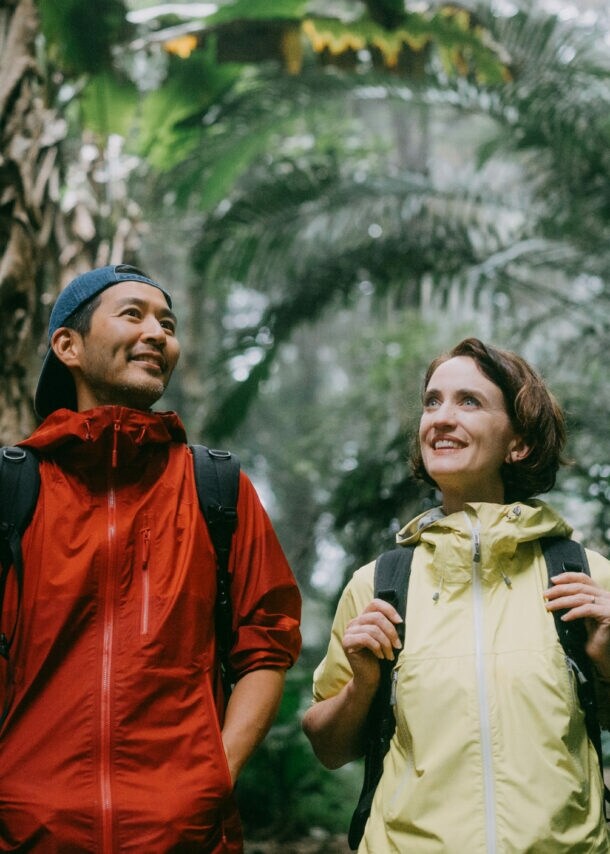 Zwei Wandernde mit Rucksäcken in einem dichten Dschungel, einer trägt eine rote Jacke, die andere eine gelbe Jacke.