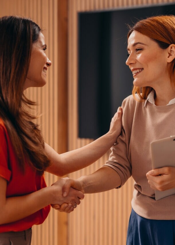Zwei Frauen geben sich die Hand und lächeln sich an. Eine Frau legt die Hand auf den Arm der anderen Frau.