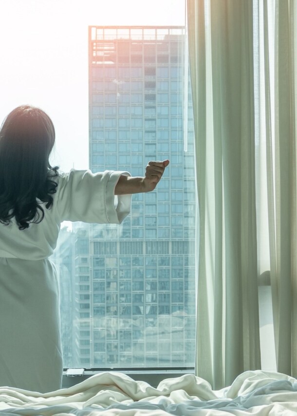 Rückansicht einer sich räkelnden Person im weißen Bademantel am Fenster eines Schlafzimmers in einer Großstadt, im Vordergrund ein Bett.