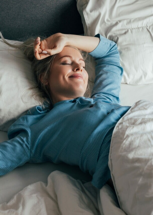 Eine zufrieden lächelnde Frau im blauen Pyjama liegt mit geschlossenen Augen in einem Bett mit weißer Bettwäsche bei Tageslicht.
