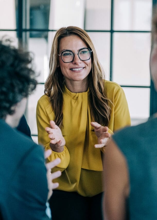 Mehrere Personen in Business-Kleidung stehen in einem Kreis, im Zentrum eine lächelnde, gestikulierende Frau im gelben Oberteil.