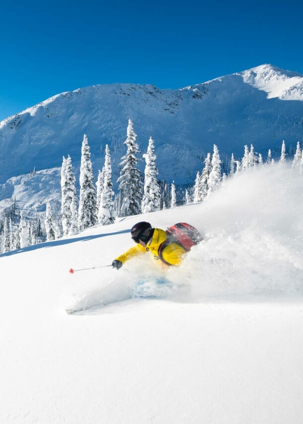 Ein Skifahrer fährt im Tiefschnee einen Hang mit Tannen hinunter vor verschneitem Bergpanorama unter blauem Himmel bei Sonnenschein.