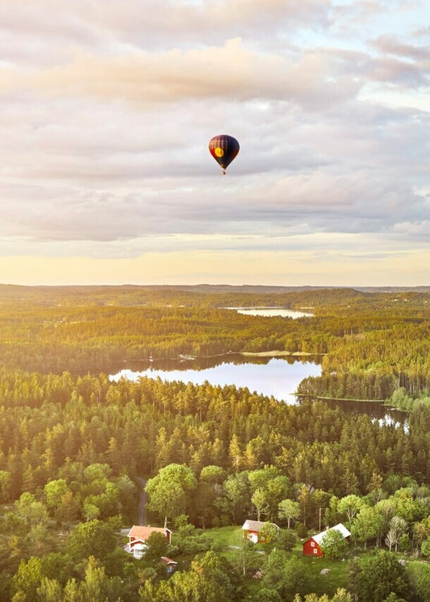 Heißluftballon mit rotem Muster fliegt über bewaldete Landschaft mit mehreren Seen und vereinzelten Häusern bei Sonnenuntergang.