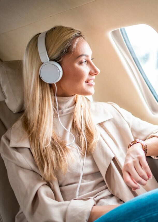 Eine Frau mit weißen Kopfhörern sitzt in einem Flugzeug in der Business-Class und blickt lächelnd aus dem Fenster.