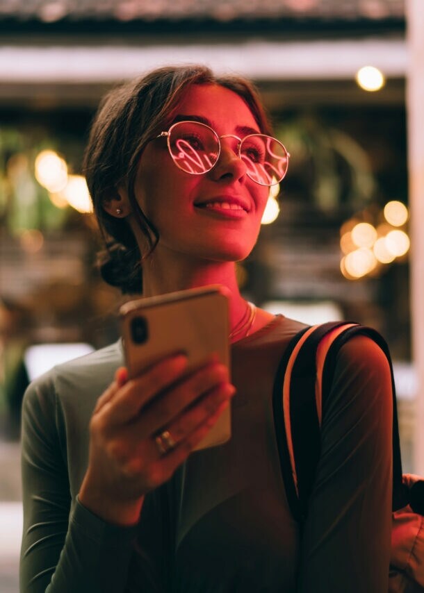 Eine junge Frau hält lächelnd ein Smartphone vor sich in der Hand, in ihrer Brille spiegelt sich der Schriftzug eines roten Logos an einer Wand.