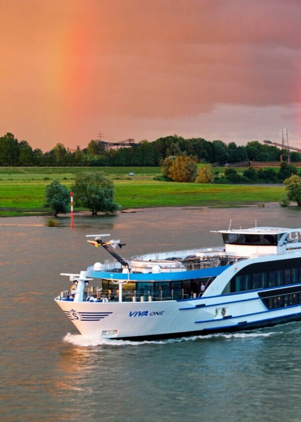 Flusskreuzfahrtschiff Viva One fährt vor grünem Flussufer mit Regenbogen am bewölkten Himmel.