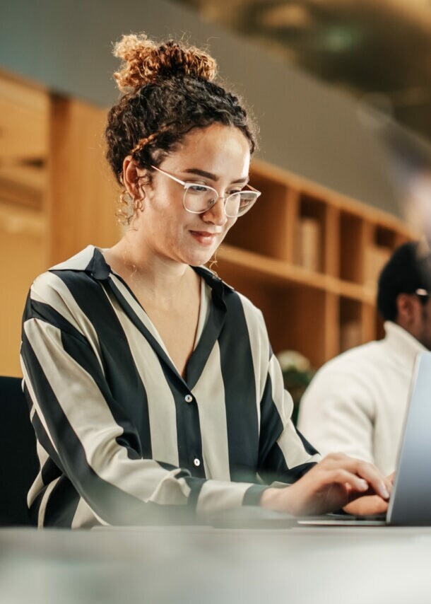 Eine junge Frau in gestreifter Bluse arbeitet an einem Laptop an einem Tisch mit weiteren Personen in einem Büro.