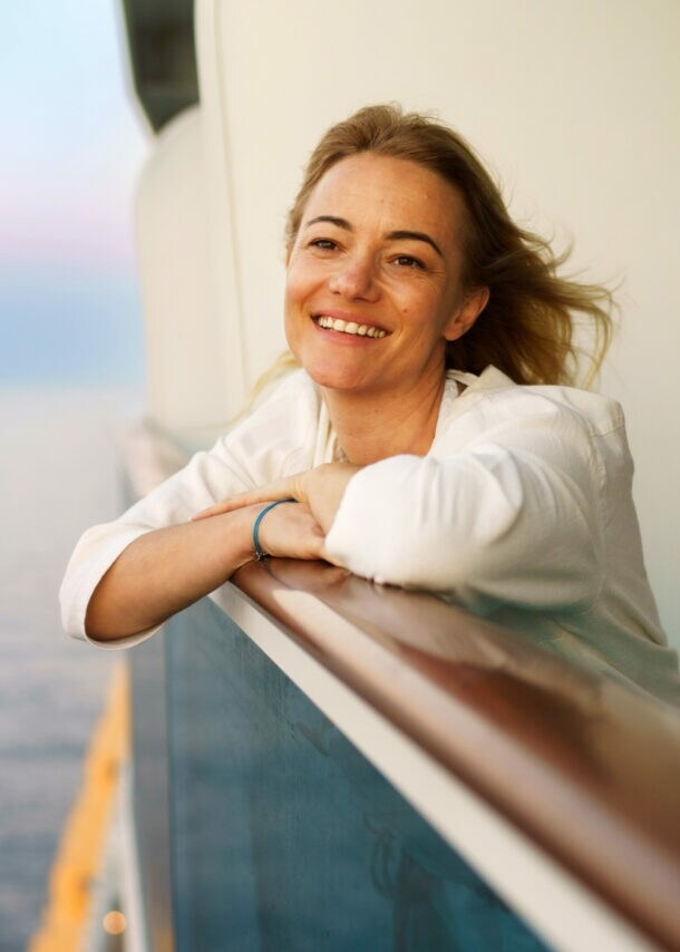 Eine lächelnde Frau lehnt an der Reling einer Balkonkabine auf einem Kreuzfahrtschiff auf See bei Abenddämmerung.