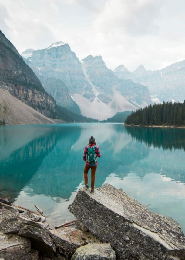Person mit rotem Rucksack steht auf Felsen am Ufer eines klaren Bergsees, umgeben von Nadelwald und hohen Bergen.